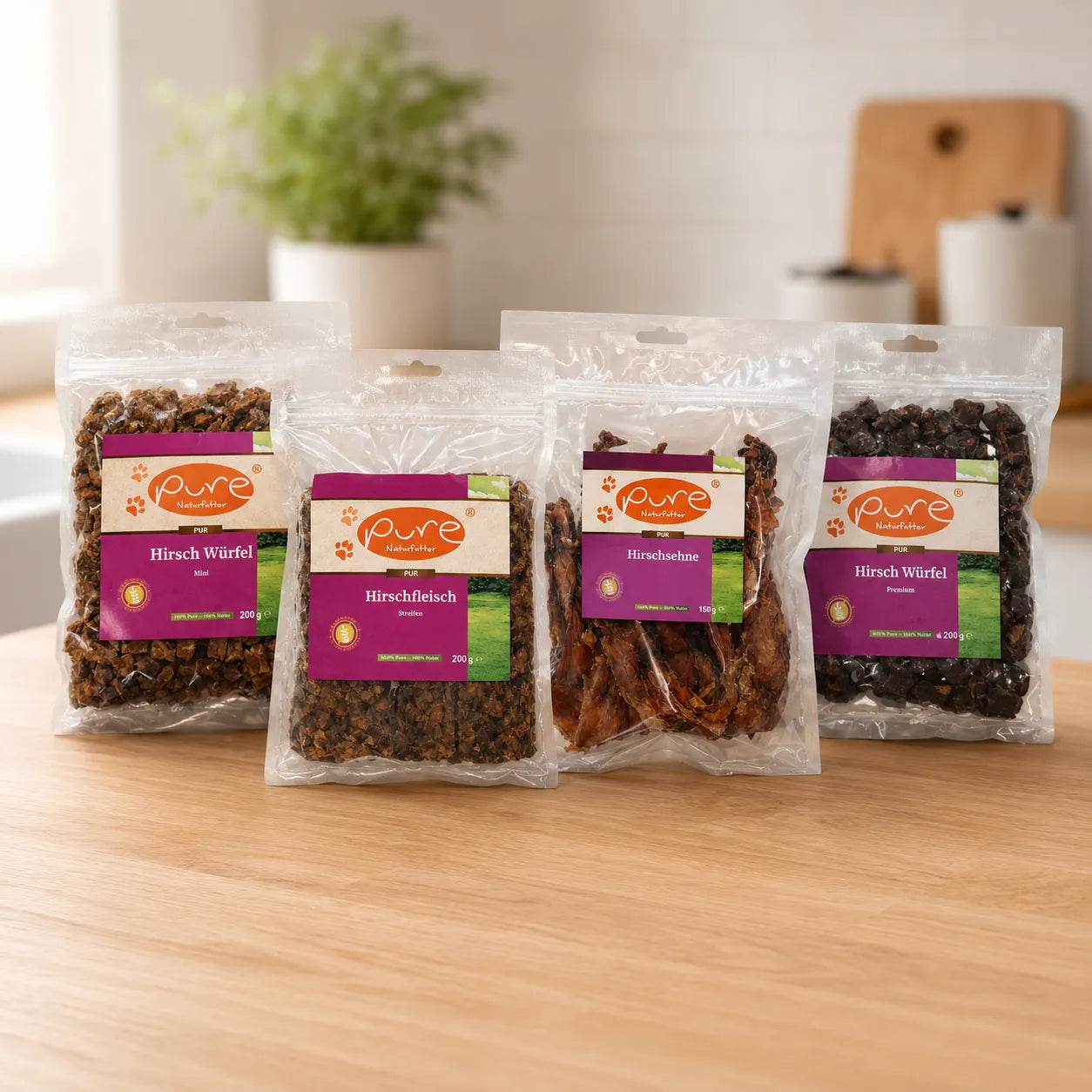 Vier PURE Naturfutter Hundesnacks mit Hirsch auf heller Küchenarbeitsplatte: Hirsch Würfel Mini, Hirschfleisch Streifen, Hirschsehne und Hirsch Würfel Premium.