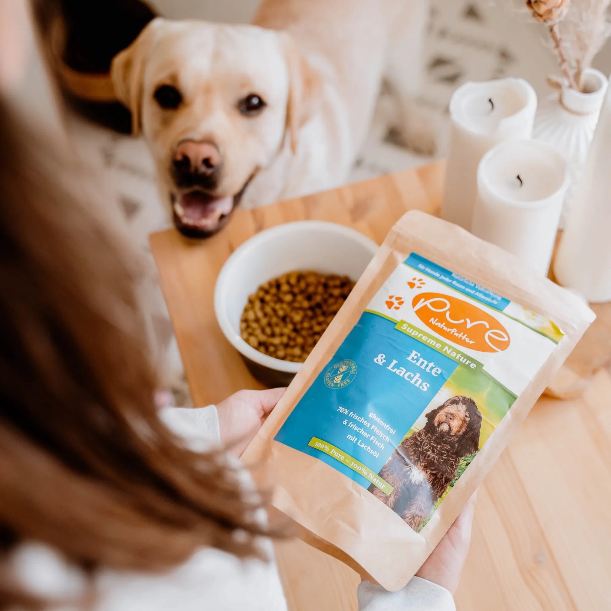Hund wartet fröhlich neben einem Napf, während ein Beutel PURE Ente & Lachs gehalten wird – hochwertiges Trockenfutter für Hunde
