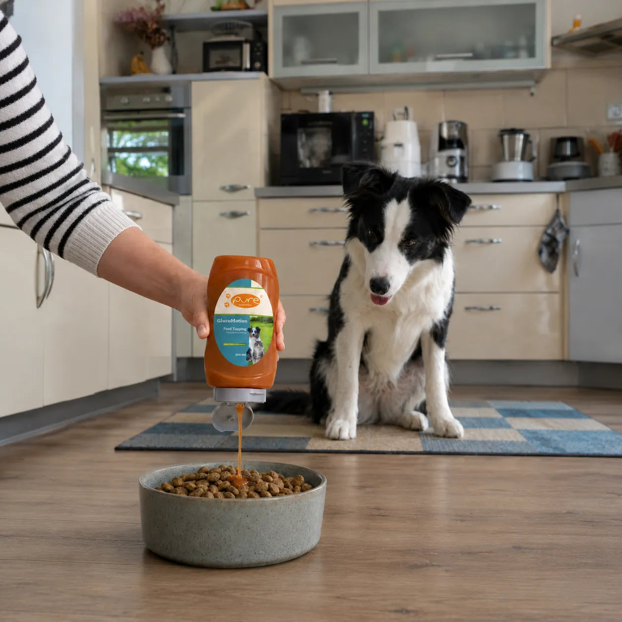 PURE GlucoMotion Food Topping wird in einer Küche über Trockenfutter gegeben, während ein schwarz-weisser Hund aufmerksam daneben sitzt