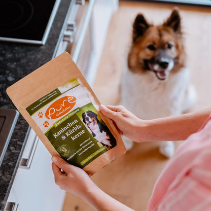 Beutel PURE Kaninchen & Kürbiskerne wird in der Küche gehalten, während ein Hund im Hintergrund sitzt
